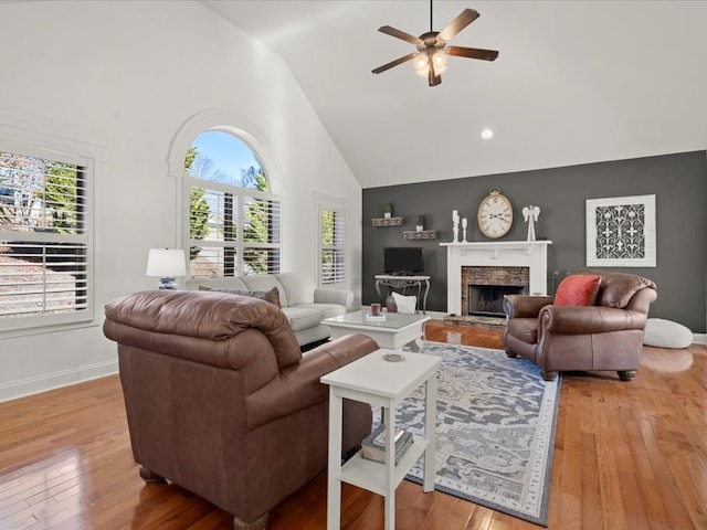 living room with ceiling fan, a stone fireplace, high vaulted ceiling, and light wood-type flooring