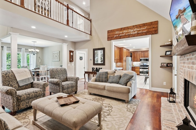 living room with high vaulted ceiling, a brick fireplace, light wood-type flooring, a notable chandelier, and decorative columns