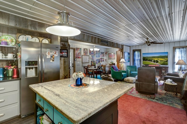 kitchen featuring stainless steel refrigerator with ice dispenser, a center island, white cabinets, ceiling fan with notable chandelier, and wood walls