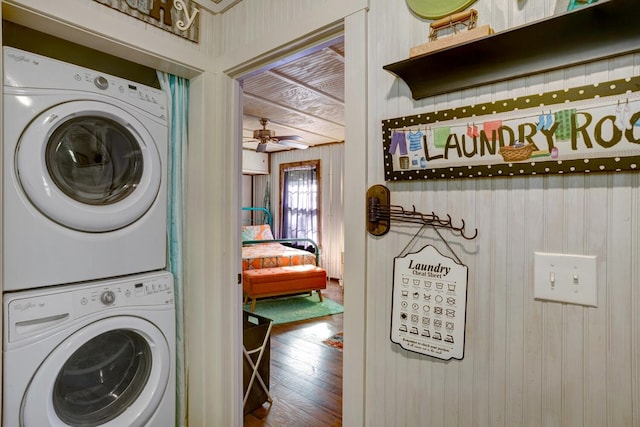 clothes washing area featuring wooden walls, stacked washer and dryer, wood-type flooring, and ceiling fan