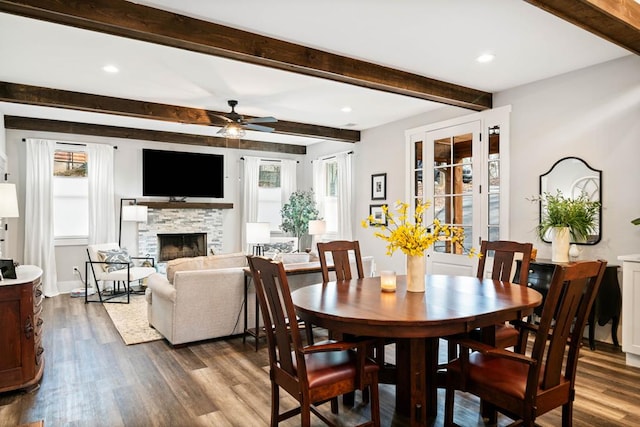 dining room featuring ceiling fan, dark hardwood / wood-style flooring, beam ceiling, and a stone fireplace