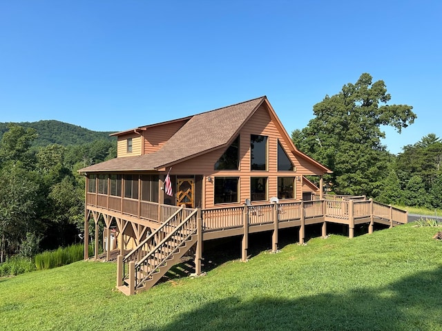 rear view of property featuring a deck, a sunroom, stairs, roof with shingles, and a lawn