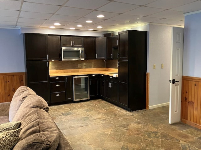 kitchen featuring tasteful backsplash, a drop ceiling, dark brown cabinets, wooden walls, and beverage cooler