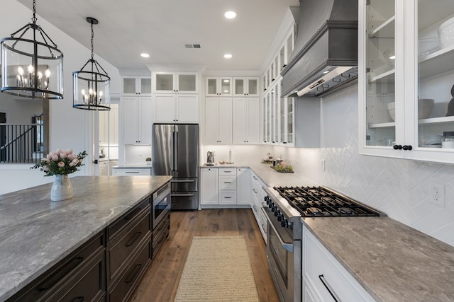 kitchen featuring high quality appliances, custom range hood, light stone countertops, a chandelier, and dark wood-style flooring