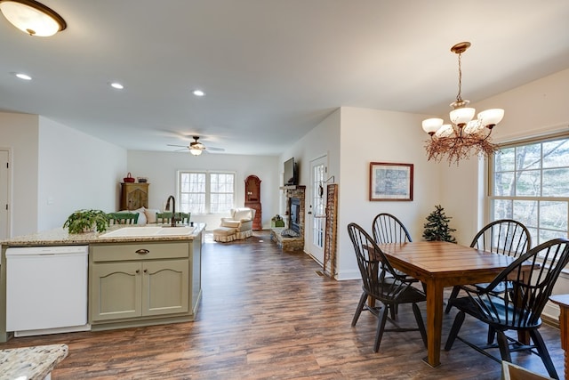 dining area featuring a stone fireplace, sink, ceiling fan with notable chandelier, and dark hardwood / wood-style flooring