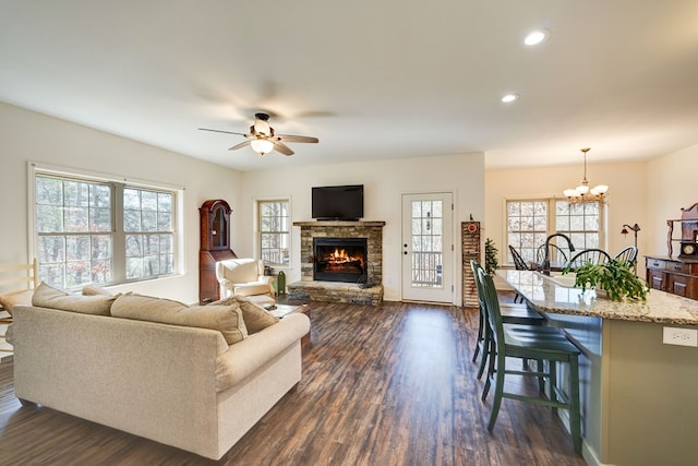 living room featuring plenty of natural light, dark wood-type flooring, ceiling fan with notable chandelier, and a stone fireplace