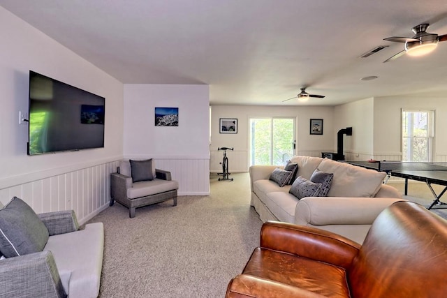 living room featuring carpet flooring, wainscoting, a wood stove, a ceiling fan, and visible vents