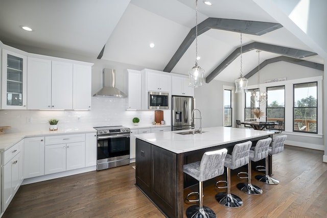 kitchen with lofted ceiling with beams, appliances with stainless steel finishes, an island with sink, wall chimney exhaust hood, and dark wood-type flooring