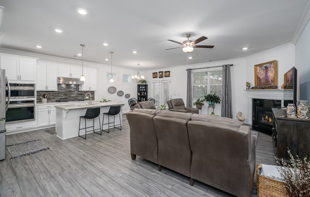 living room with ceiling fan with notable chandelier, light wood-type flooring, crown molding, and sink