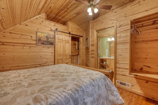 bedroom featuring wood-type flooring, ensuite bath, vaulted ceiling, a barn door, and wood walls