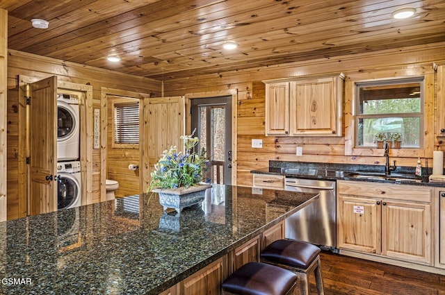 kitchen with dishwasher, sink, stacked washer and dryer, wooden walls, and dark stone countertops