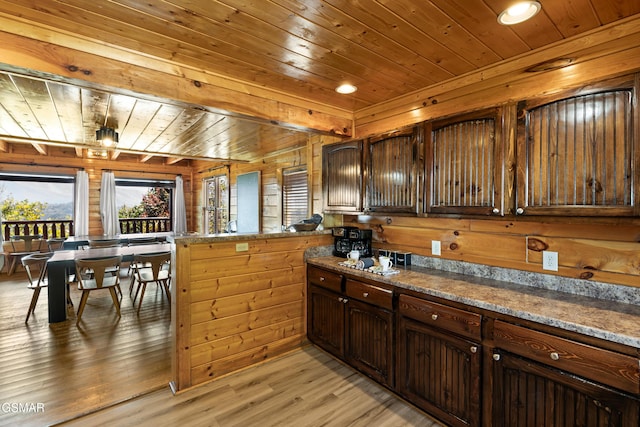 kitchen featuring kitchen peninsula, dark brown cabinets, light wood-type flooring, dark stone countertops, and wooden walls