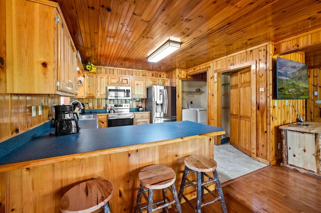 kitchen with stainless steel appliances, wood walls, light hardwood / wood-style floors, a kitchen breakfast bar, and washer and clothes dryer