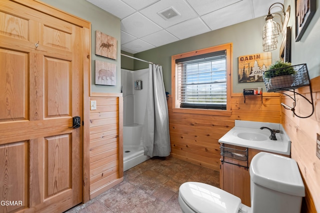 bathroom featuring a drop ceiling, vanity, wooden walls, toilet, and walk in shower