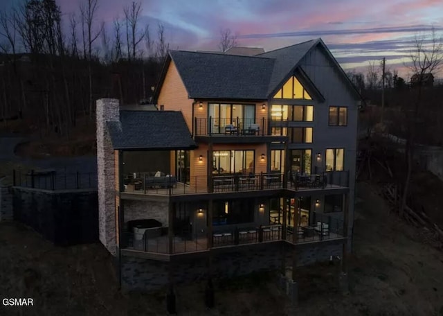 back of house at dusk with roof with shingles, a chimney, and a balcony