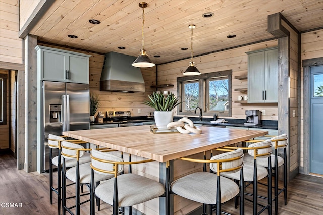 kitchen with wooden ceiling, stainless steel appliances, wood walls, wooden counters, and wall chimney exhaust hood