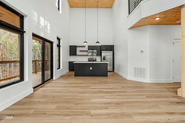 kitchen with stainless steel appliances, a towering ceiling, hanging light fixtures, a center island with sink, and wood ceiling