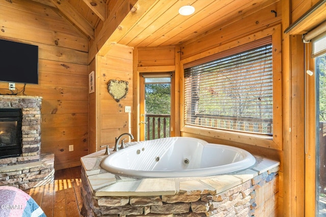 bathroom with vaulted ceiling, hardwood / wood-style flooring, wood ceiling, a stone fireplace, and a relaxing tiled tub