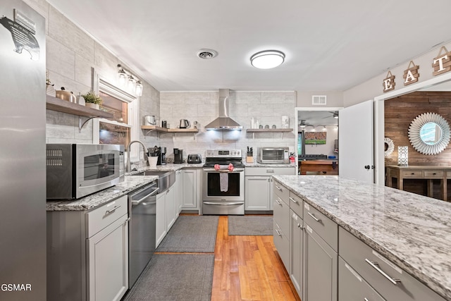 kitchen featuring sink, wall chimney exhaust hood, light wood-type flooring, appliances with stainless steel finishes, and light stone counters