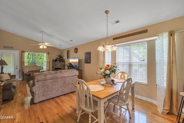 dining space with light wood-type flooring, a stone fireplace, vaulted ceiling, a chandelier, and ceiling fan