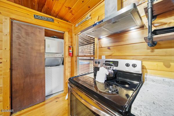 kitchen featuring stainless steel range with electric cooktop, wood ceiling, wooden walls, stacked washer / dryer, and wall chimney range hood