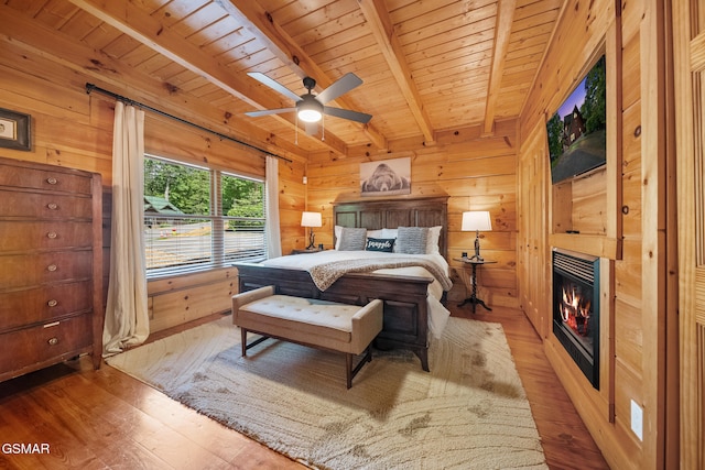 bedroom with light wood-style floors, wooden ceiling, beam ceiling, wood walls, and a warm lit fireplace