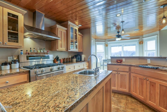 kitchen featuring a kitchen island with sink, a sink, wall chimney exhaust hood, light stone countertops, and stainless steel range with gas stovetop