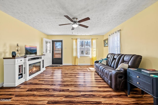 living area with baseboards, wood finished floors, a glass covered fireplace, a textured ceiling, and a ceiling fan