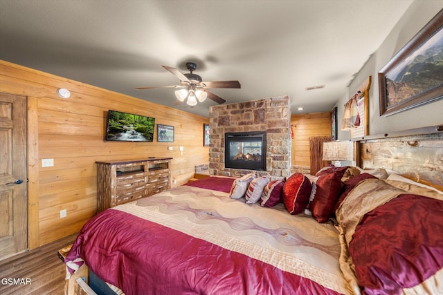 bedroom featuring a stone fireplace, wood walls, wood finished floors, visible vents, and a ceiling fan