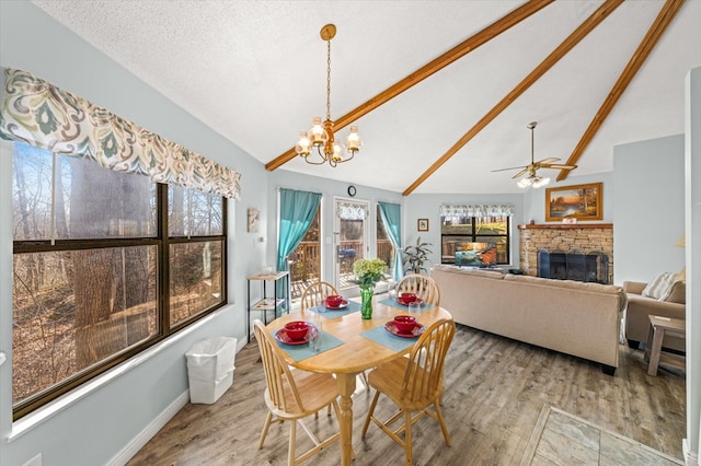 dining area featuring light wood-type flooring, a textured ceiling, ceiling fan with notable chandelier, lofted ceiling with beams, and a stone fireplace