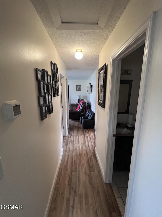 hall featuring baseboards, a textured ceiling, attic access, and wood finished floors