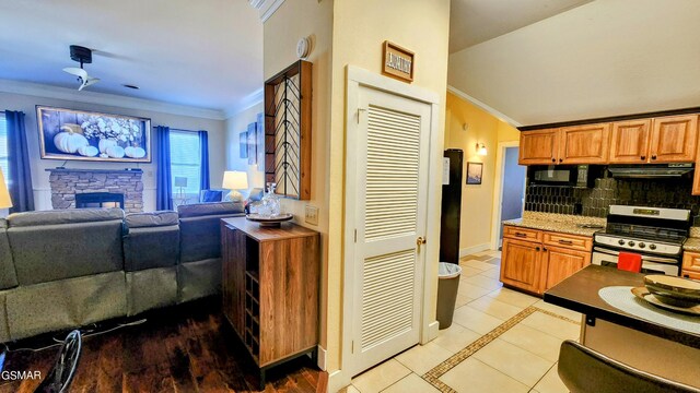 kitchen featuring stainless steel gas stove, backsplash, ornamental molding, a stone fireplace, and exhaust hood