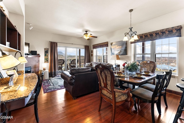 dining room featuring dark wood finished floors, ceiling fan with notable chandelier, a fireplace, and baseboards