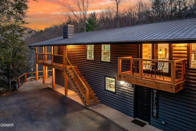 back of house at dusk featuring a balcony, faux log siding, a chimney, metal roof, and stairway