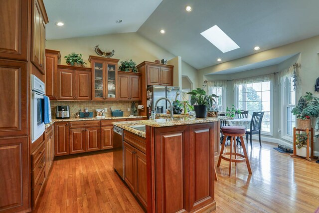 kitchen featuring light stone countertops, stainless steel appliances, tasteful backsplash, an island with sink, and vaulted ceiling with skylight