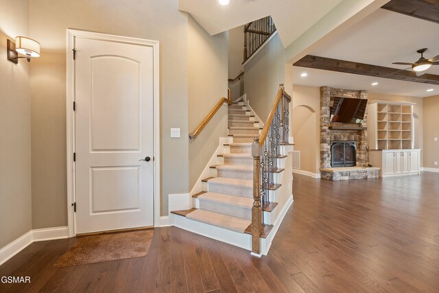 staircase with beam ceiling, a stone fireplace, ceiling fan, and wood-type flooring