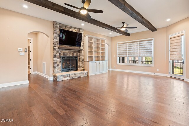 unfurnished living room featuring ceiling fan, beam ceiling, a stone fireplace, and wood-type flooring