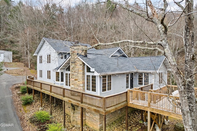 rear view of property featuring a deck, stone siding, roof with shingles, and a chimney