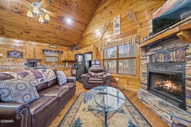 living room with a stone fireplace, ceiling fan, wooden walls, light wood-style flooring, and high vaulted ceiling