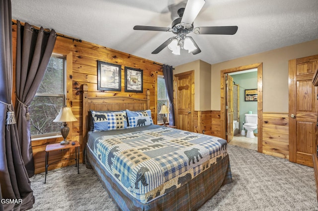 carpeted bedroom with a textured ceiling, ensuite bath, a ceiling fan, wood walls, and wainscoting