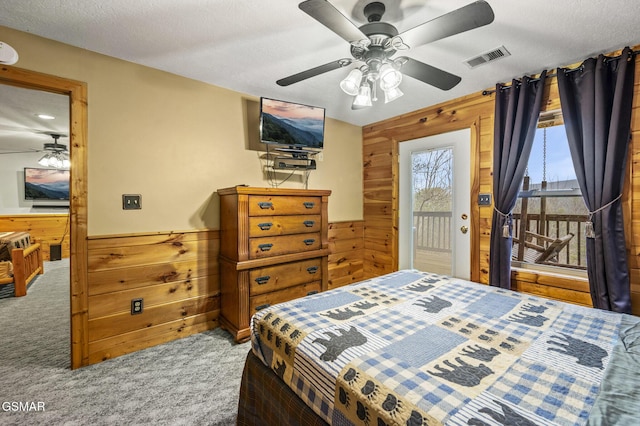 carpeted bedroom with a textured ceiling, a ceiling fan, a wainscoted wall, wood walls, and visible vents