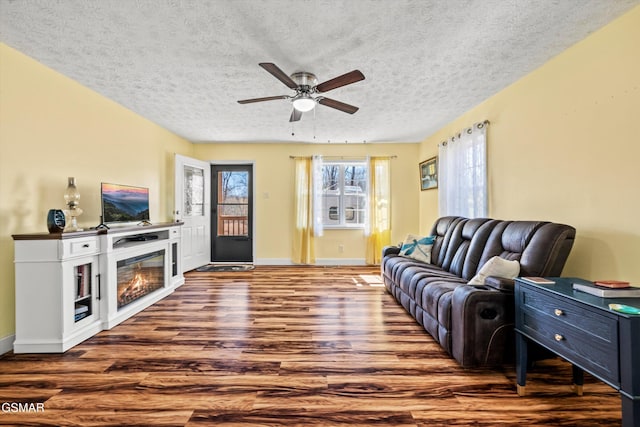 living area featuring ceiling fan, a textured ceiling, wood finished floors, baseboards, and a glass covered fireplace