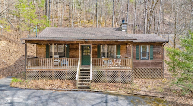 view of front of house featuring a shingled roof, a porch, a forest view, and a chimney