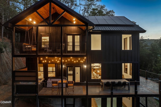 back of house at dusk with french doors, board and batten siding, roof mounted solar panels, metal roof, and a balcony