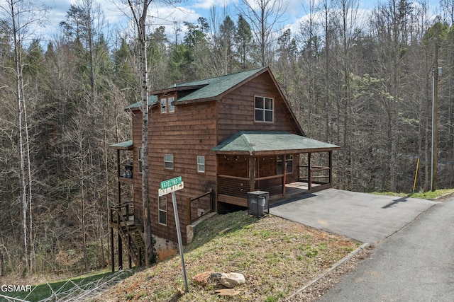 view of property exterior with covered porch, a forest view, a shingled roof, and driveway