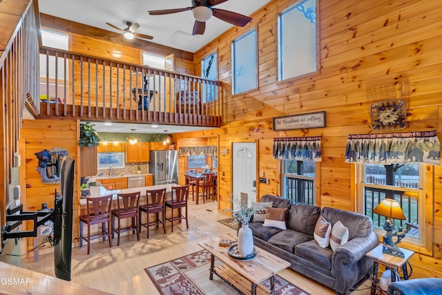 living room with ceiling fan, a towering ceiling, light wood-type flooring, and wood walls