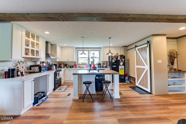 kitchen featuring black appliances, wall chimney exhaust hood, a barn door, light wood-style flooring, and white cabinets