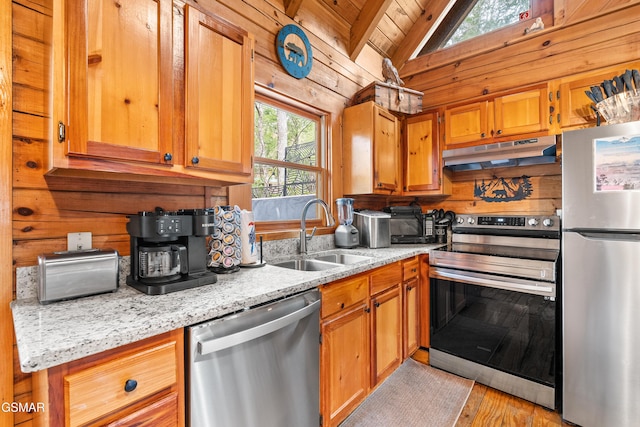 kitchen featuring wooden walls, under cabinet range hood, vaulted ceiling, appliances with stainless steel finishes, and a sink