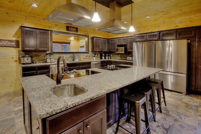 kitchen with stainless steel appliances, light stone counters, wood walls, an island with sink, and exhaust hood