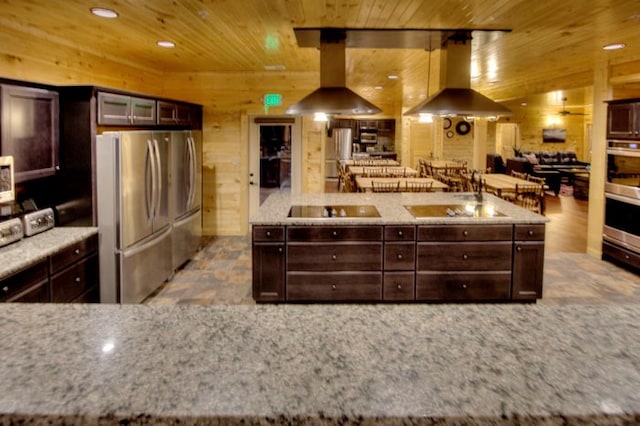 kitchen with wood walls, black electric cooktop, island exhaust hood, and stainless steel refrigerator